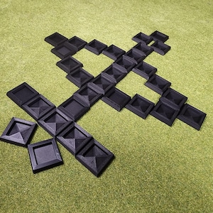 May include: Arrangement of black, square-shaped objects with pyramidal tops, forming a cross-like design on a green surface. The objects are evenly spaced, creating a geometric pattern. The overall aesthetic is minimalist and modern.