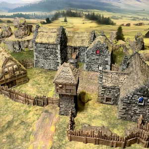 May include: A miniature tabletop gaming village set featuring several stone buildings with thatched roofs, a wooden watchtower, and a surrounding wooden fence. The structures are arranged in a cluster on a green landscape mat with painted hills.