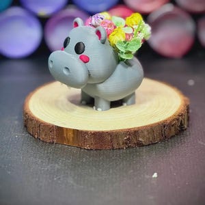 May include: A gray 3D-printed hippo planter with a small bouquet of colorful flowers. The hippo has black eyes, pink cheeks, and a small mouth. It sits on a round wooden slice.