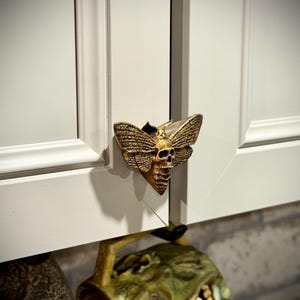 May include: A gold-colored cabinet pull featuring a moth design with a skull face. The pull is mounted on a white cabinet door. A zombie-themed mug hangs below the pull.