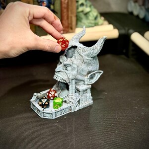 May include: A gray, horned demon head dice holder with a detailed, weathered appearance. A red and white die is being placed into the head. Other dice in various colors are in the tray below. The dice holder is designed for tabletop gaming.