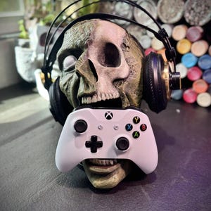 May include: A zombie skull wearing black headphones and holding a white Xbox controller. The skull is green and brown with a detailed, weathered texture. The controller has colored buttons and the Xbox logo.