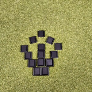May include: A collection of small, square, black objects arranged in a geometric pattern on a green surface. The objects appear to be made of a textured material, possibly plastic or rubber. The arrangement suggests a puzzle or game component.