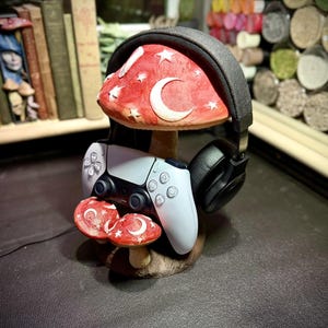 May include: A whimsical mushroom-shaped headphone stand with a red cap adorned with white stars and a crescent moon. A white game controller rests below the mushroom cap. Black headphones are placed on the stand.