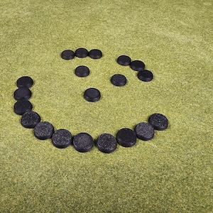 May include: A collection of small, black, circular objects arranged in a smiley face pattern on a green surface. The objects appear to be made of a textured material. The arrangement creates a playful and simple design.