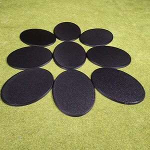 May include: A collection of nine black oval-shaped bases, likely for miniatures or models, arranged on a green surface. The bases have a textured surface and are of varying sizes. The background is a solid green color.