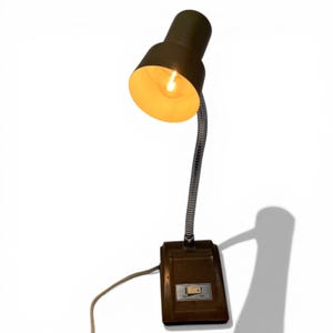 May include: A vintage desk lamp with a brown base and shade, featuring a flexible, silver-colored neck. The lamp is turned on, emitting a warm, yellow light. The base has an on/off switch and a power cord.