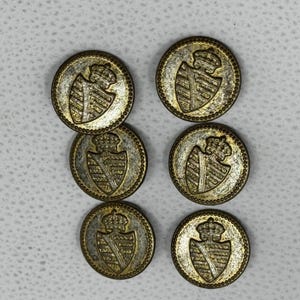 May include: A set of six circular brass-toned metal buttons, each featuring an embossed heraldic shield design topped with a crown. The buttons have a textured, aged finish with a beaded border detail around the outer edge of each piece.