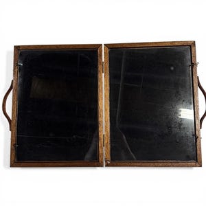 May include: A folding wooden framed mirror with two panels. The mirror has a dark surface and is framed in a light brown wood. Brown leather handles are attached to the sides. The mirror is designed to fold in half.