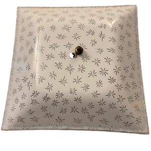 May include: Square, frosted glass ceiling light fixture with a starburst pattern. The fixture has a central mounting hardware with a silver and brass finish. The edges of the glass are slightly textured.