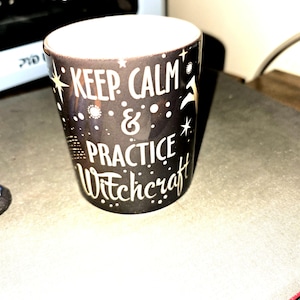 May include: A black shot glass with the words "KEEP CALM & PRACTICE Witchcraft" in white lettering. The glass has a white interior and is decorated with stars and dots. The shot glass is cylindrical and appears to be made of ceramic.