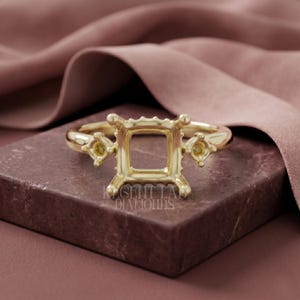 May include: A gold-colored ring setting with a square center and two smaller side settings. The ring is displayed on a dark, textured surface with the text "REGENT LAB DIAMONDS" visible. The background features a draped, pink fabric.