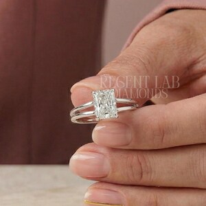 Könnte beinhalten: Ein silberner Ring mit einem rechteckigen Diamanten im Radiant-Schliff. Der Ring hat ein geteiltes Schaftdesign. Der Diamant wird von Krappen gehalten. Der Text "REGENT LAB DIAMONDS" ist im Hintergrund sichtbar. Ein klassisches Schmuckstück.