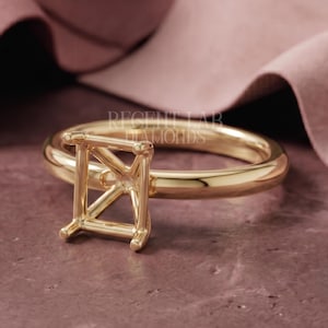 May include: A gold ring setting featuring a rectangular four-prong head designed to hold a gemstone. The band is smooth and polished. The text RECENT LAB DIAMONDS is visible in the center of the ring setting, which is positioned on a textured surface.