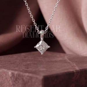 May include: A silver-toned necklace featuring a princess-cut diamond pendant suspended from a delicate chain. The text REGENT LAB DIAMONDS is visible in the background. The pendant is set in a four-prong metal setting, showcasing a clear, faceted stone.