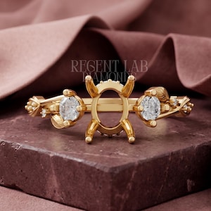 May include: A gold ring setting featuring a central oval prong mount, flanked by two round brilliant-cut diamonds. The band is decorated with small leaf motifs and additional tiny accent stones. The text REGENT LAB DIAMONDS is visible.