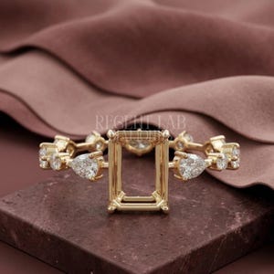 May include: A gold ring setting featuring a central rectangular prong mount for a stone, flanked by alternating round and pear-shaped clear stones along the band. The text REGENT LAB is visible in the center of the ring setting.
