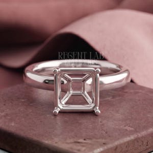 May include: A polished silver-toned metal ring setting featuring a square-shaped four-prong head designed to hold a gemstone. The band is smooth and rounded, and the text REGENT LAB DIAMONDS is visible in the background behind the ring setting.