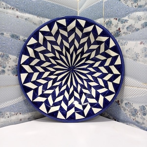 May include: A large, round ceramic serving bowl with a striking geometric design. The bowl features a deep blue rim and a central pattern of white and blue diamond shapes radiating outwards. This decorative bowl is perfect for serving salads or as a centerpiece.