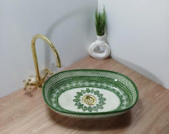 May include: Oval ceramic sink with a green and white floral design. The sink has a gold-colored drain and is set on a wooden surface. A gold faucet and a white vase with greenery are also visible.