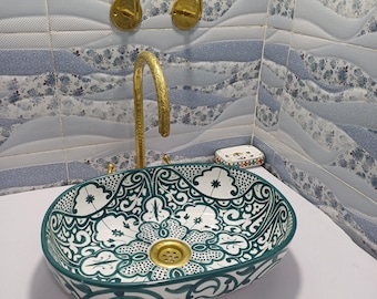 May include: An oval ceramic sink with a white and teal floral design. The sink has a gold-colored drain and is mounted on a white surface. A gold faucet and two gold hooks are mounted on the wall behind the sink. The wall is decorated with blue and white floral tiles.