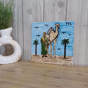 May include: A square ceramic tile featuring a hand-painted scene of a person in a green robe and yellow head covering standing next to a camel in a desert landscape. Two palm trees, birds in a blue sky, and the word FES are painted on the tile.