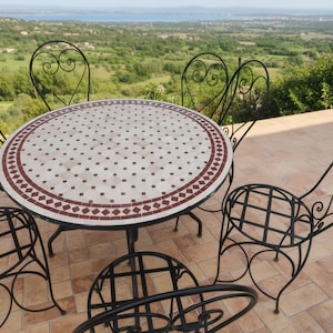Peut inclure: Une table de jardin ronde avec un dessus en mosaïque crème, rouge et marron. La table est entourée de quatre chaises en fer forgé noir avec des volutes décoratives. L'ensemble est sur un patio carrelé avec une vue panoramique.