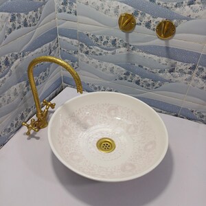 May include: A white oval bathroom sink with a floral pattern, paired with a gold faucet and drain. The sink is set against a backdrop of blue and white patterned tiles and two gold wall fixtures.