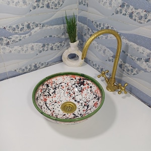 Pode incluir: Uma pia de cerâmica decorativa com interior branco, com design de tinta salpicada preta e vermelha. A pia tem uma borda verde e um ralo de latão. Uma torneira dourada e um pequeno vaso com vegetação também são visíveis.