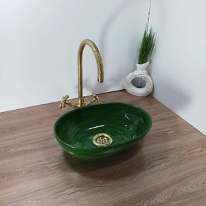 Könnte beinhalten: Ovales, smaragdgrünes Keramikwaschbecken mit Messingablauf und Wasserhahn. Der Wasserhahn hat einen gebogenen Auslauf und Kreuzgriffe. Eine weiße Vase mit kreisförmigem Design hält Grün, auf einer Holzoberfläche.