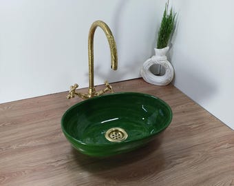 May include: Oval-shaped, emerald green ceramic sink with a brass drain and faucet. The faucet has a curved spout and cross handles. A white vase with a circular design holds greenery, set on a wooden surface.