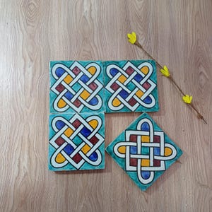 Op de afbeelding: Vier vierkante keramische tegels met een turquoise rand en een kleurrijk Keltisch knoopontwerp in blauw, geel en rood. Eén tegel is schuin en een takje gele bloemen staat op de achtergrond.