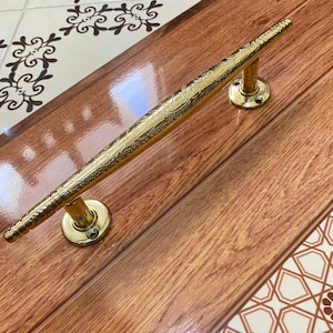 May include: A gold-colored door pull with an ornate, textured design. The handle is mounted on two circular bases with screw holes. The door pull is set against a wood-grain surface.