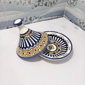Może przedstawiać: Ceramiczny tagine z stożkową pokrywą i pasującym talerzem. Tagine ma białą podstawę z niebieskimi, żółtymi, czerwonymi i zielonymi geometrycznymi wzorami. Pokrywa jest otwarta.