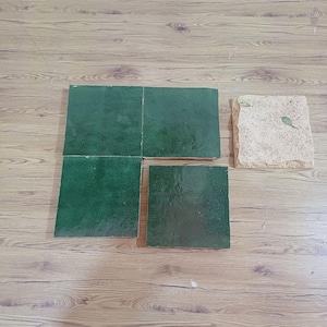 Op de afbeelding: Vier vierkante tegels worden getoond. Drie tegels hebben een glanzende, diepgroene afwerking met subtiele oppervlaktevariaties. Eén tegel is apart geplaatst en toont een ruw, ongeglazuurd, lichtbeige getextureerd oppervlak aan de bovenzijde.