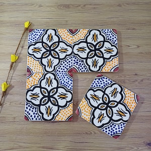 Op de afbeelding: Handgeschilderde keramische tegels met een bloemmotief. De tegels hebben een witte bloem met zwarte contouren, gele accenten en blauwe en oranje gestippelde achtergronden. De randen zijn rood.