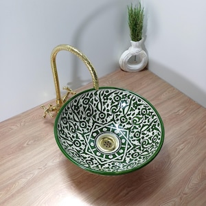 May include: A round ceramic sink with a green and white floral design. It features a gold tap and a green border. The sink is placed on a wooden surface. A small white vase with greenery is in the background. The sink's diameter is approximately 40 cm.
