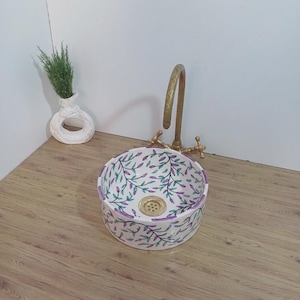 以下が含まれることがあります： 紫と緑の花柄が施された、丸い白いセラミック洗面台。洗面台には金色の排水口があり、ヴィンテージスタイルの真鍮製蛇口と組み合わされています。グリーンの入った小さな白い花瓶が横に置かれています。