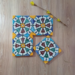 Op de afbeelding: Vier vierkante keramische tegels met een kleurrijk bloemmotief in blauw, groen, rood en geel. De tegels zijn gerangschikt op een houten oppervlak en tonen een decoratief patroon. Een gele bloemstengel is rechtsboven geplaatst.
