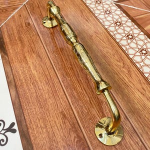 Puede incluir: Un tirador de puerta dorado con detalles florales ornamentados. El mango es curvo y se fija a la puerta con placas circulares. El tirador de la puerta es de metal y tiene un acabado brillante.