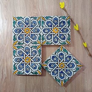 Puede incluir: Cuatro baldosas de cerámica cuadradas con un diseño geométrico en azul, blanco, amarillo y verde. Las baldosas están dispuestas sobre una superficie de madera, con una ramita de flores amarillas. Las baldosas presentan un patrón intrincado y repetitivo.