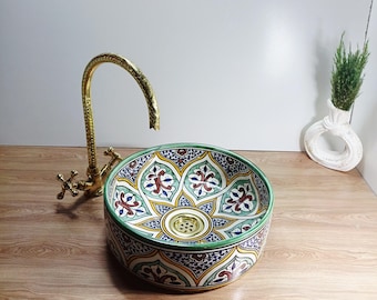 May include: A decorative ceramic sink with a colorful floral pattern in shades of blue, green, red, and yellow. The sink is round and sits on a wooden surface. A gold-colored faucet is positioned to the left of the sink.