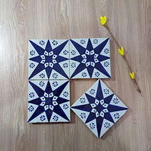 Puede incluir: Cuatro baldosas de cerámica blancas con un diseño de estrella azul. Las baldosas están dispuestas sobre una superficie de madera, con una ramita de flores amarillas. El patrón de estrella está compuesto por formas geométricas y acentos florales.