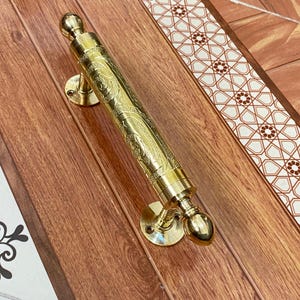 May include: A gold-colored door handle with intricate engraved floral patterns. The handle has a cylindrical bar with rounded ends and circular mounting plates. The handle is set against a wood-grain surface with decorative tile borders.