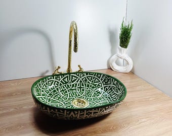 May include: An oval-shaped, green and white patterned ceramic sink with a gold faucet. The sink has a decorative design and is set on a wooden surface. A small white vase with greenery is in the background.