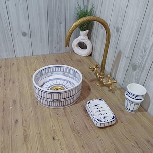 May include: A round ceramic vessel sink with blue and white striped patterns, a brass faucet with two cross handles, a rectangular soap dish with blue accents, and a matching cup, all featuring hand-painted blue line and dot decorative motifs.