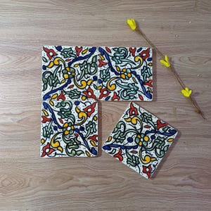 May include: Four ceramic tiles with a floral design in red, yellow, green, and blue on a white base. The tiles are arranged on a wooden surface, with a sprig of yellow flowers in the background.