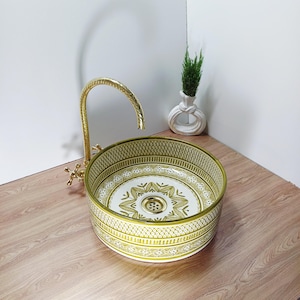 Op de afbeelding: Een ronde keramische wastafel met een gouden kraan. De wastafel heeft een wit interieur met een gouden bloemenpatroon en een gouden rand. De kraan en de afvoer zijn ook goudkleurig. Een kleine witte vaas met een groene plant staat op de achtergrond.