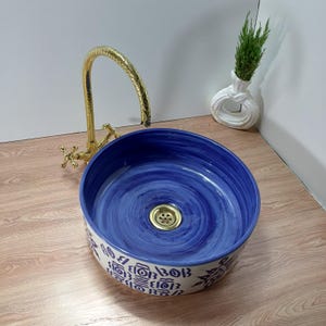 May include: A round, blue and white ceramic sink with a gold faucet. The sink has a blue interior with a swirl pattern and a decorative border. A small white vase with greenery is in the background.