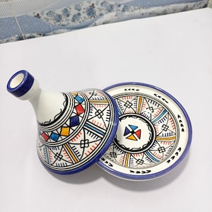 Puede incluir: Un tajine de cerámica blanco y azul con tapa, con coloridos patrones geométricos en rojo, amarillo, azul y negro. El tajine está abierto, revelando el interior. La base también está decorada con patrones similares.